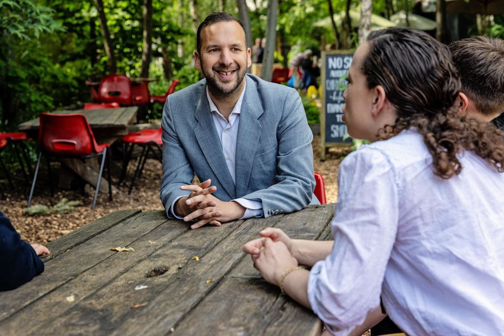 Zack Polanski speaks with Londoners while seated at a wooden picnic table in a community garden. 