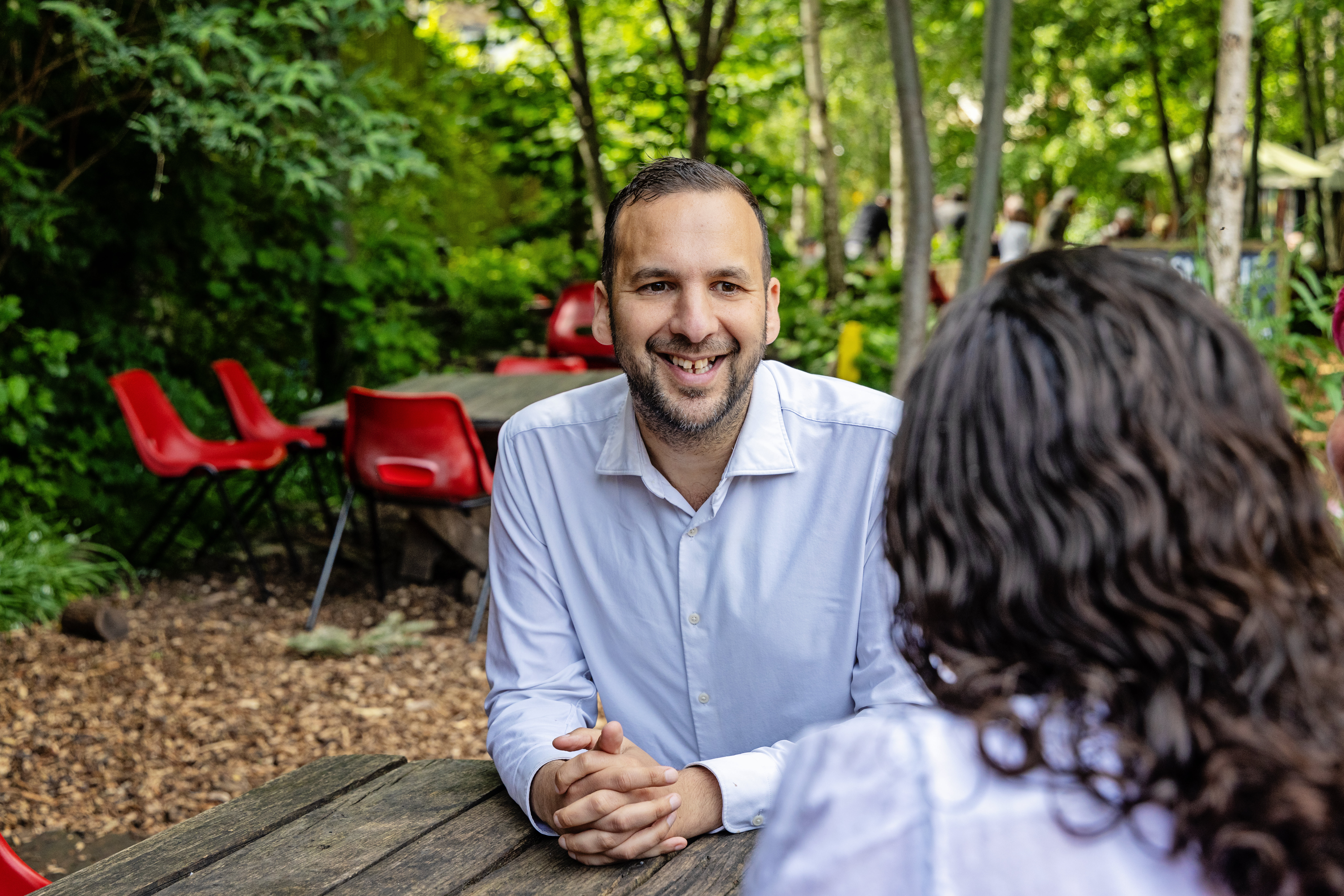 Zack Polanski speaks with Londoners while seated at a wooden picnic table in a community garden. 