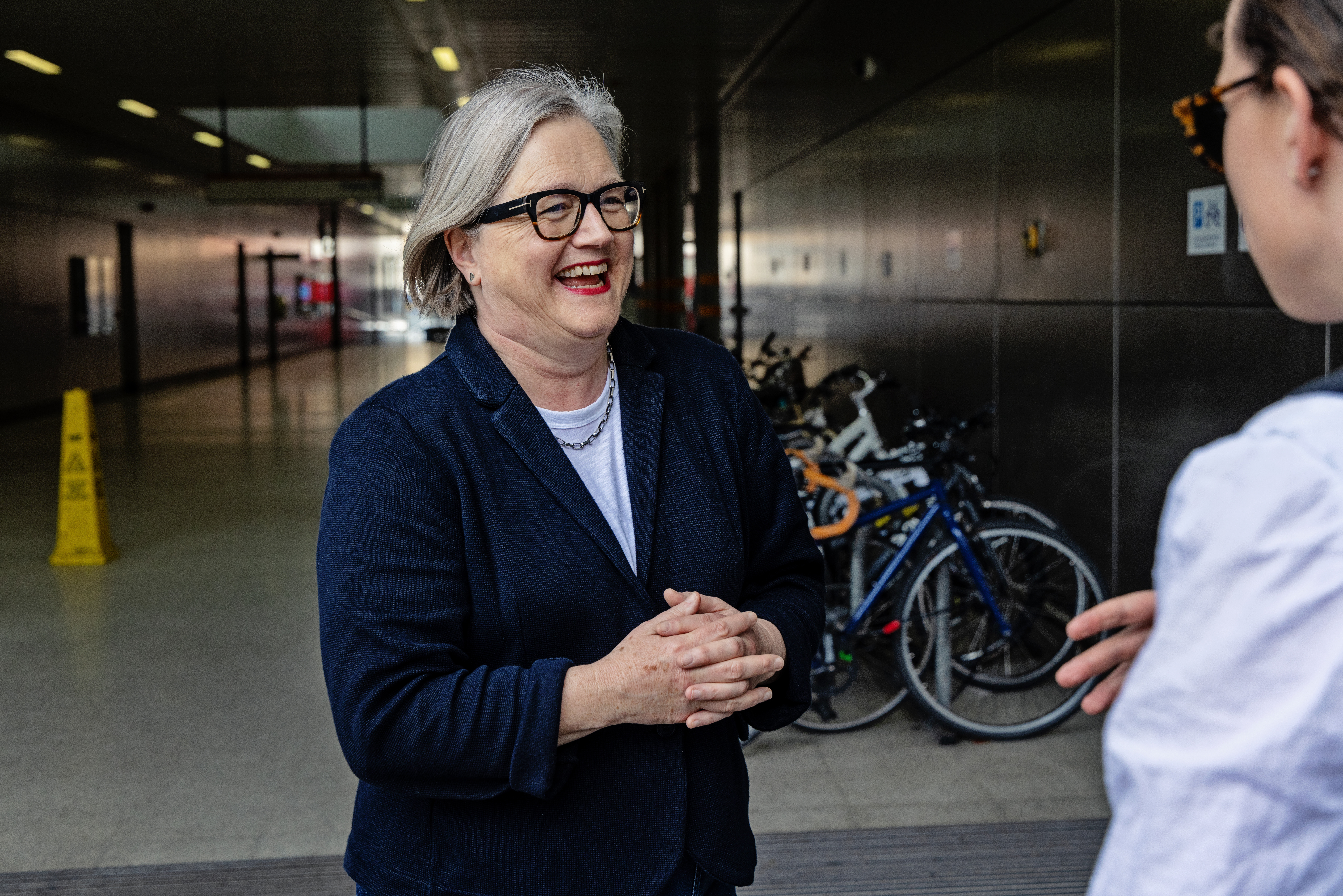 Caroline Russell AM laughs off to the side of an image, standing in front of bike racks at a London transport hub.