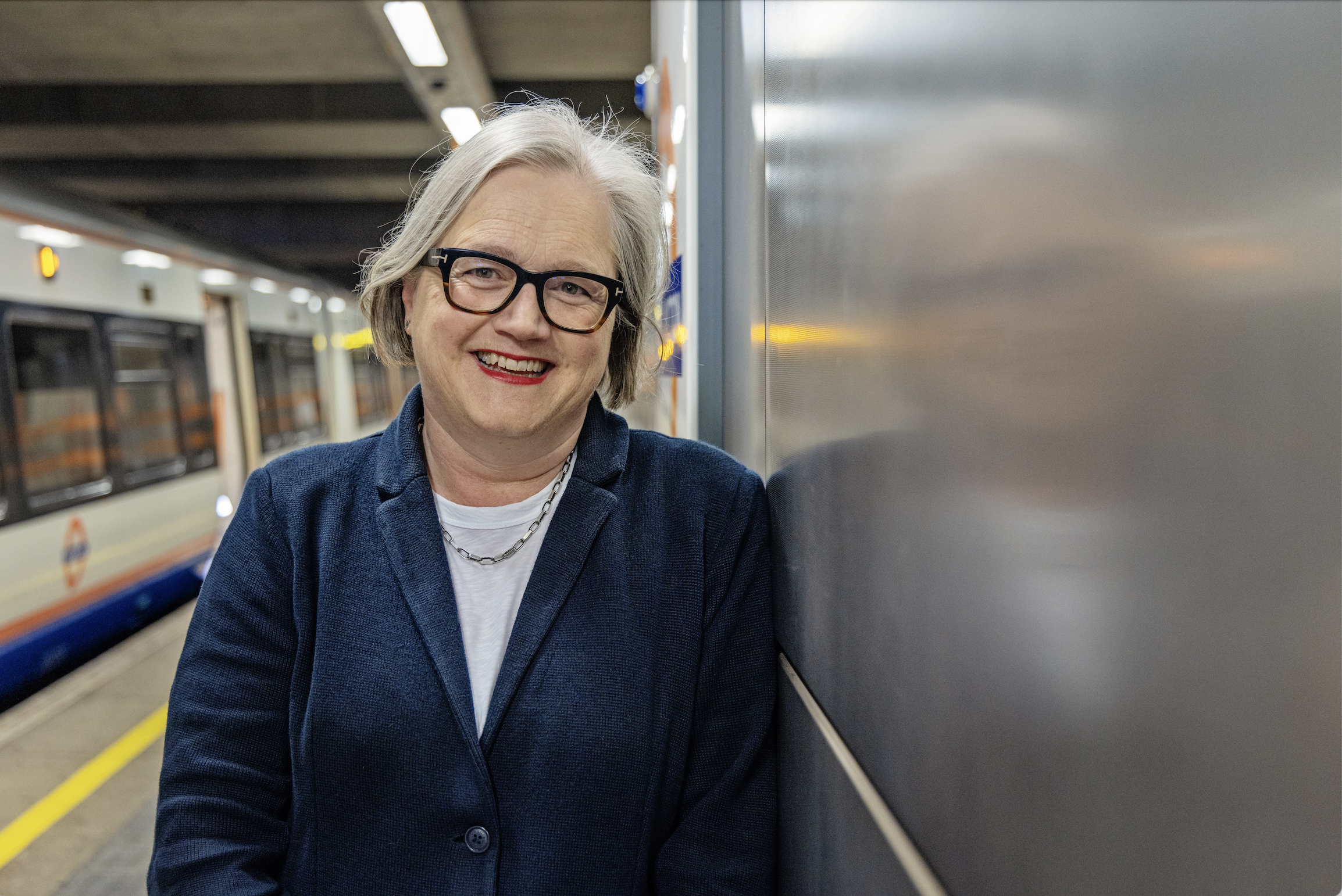 Caroline Russell AM laughs off to the side of an image, standing in front of bike racks at a London transport hub.